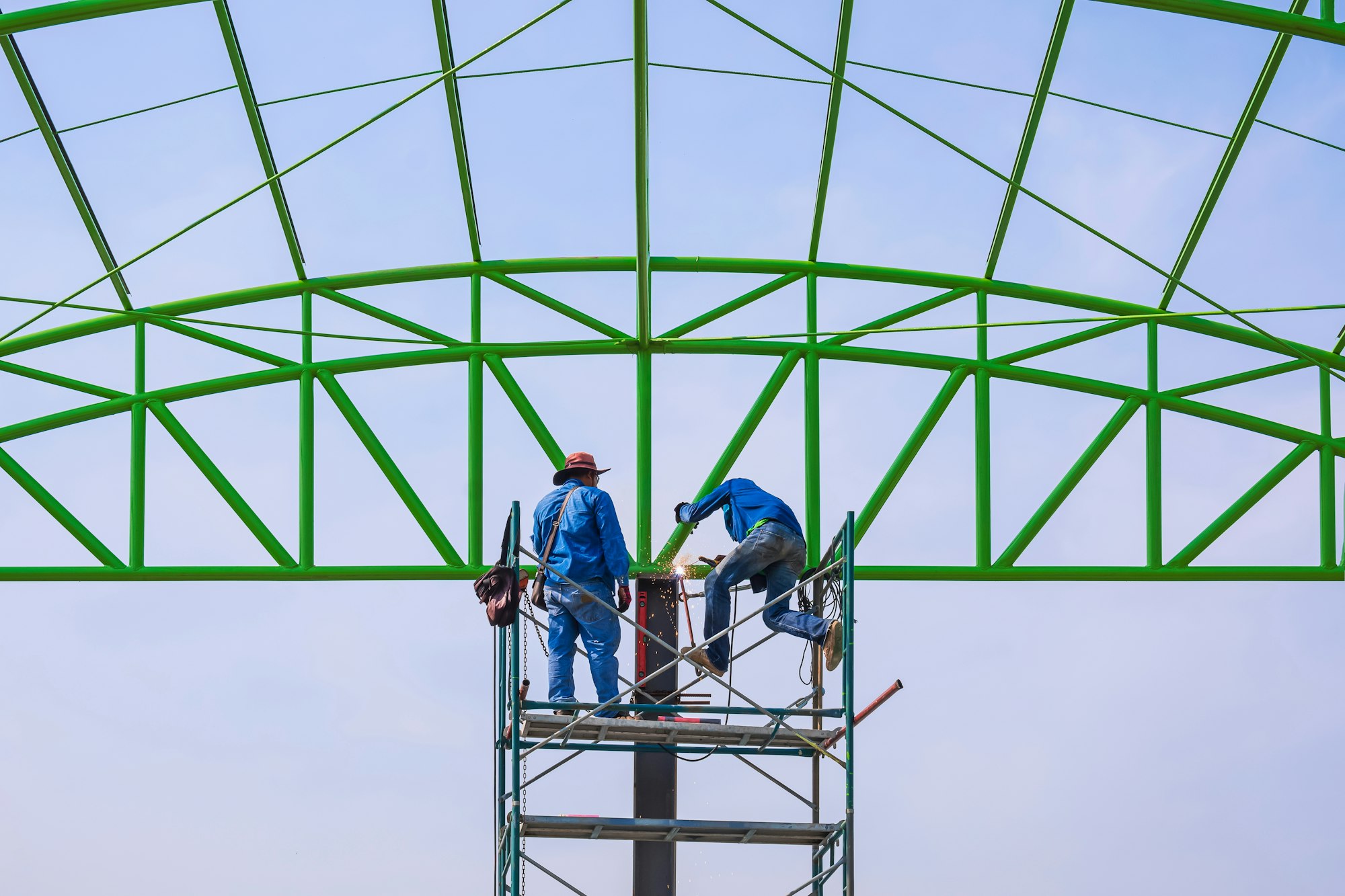 Two construction workers on scaffolding working to build metal roof structure of industrial building